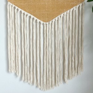 May include: A woven wall hanging with a triangular, golden-yellow top section and long, off-white fringe. The fringe hangs in vertical lines, creating a textured, bohemian aesthetic. The wall decor is designed to add a touch of warmth and style to any room.