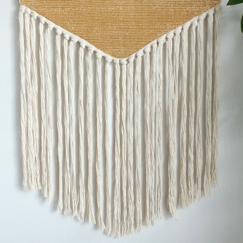 May include: A woven wall hanging with a triangular, golden-yellow top section and long, off-white fringe. The fringe hangs in vertical lines, creating a textured, bohemian aesthetic. The wall decor is designed to add a touch of warmth and style to any room.