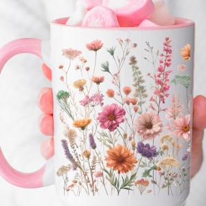 May include: White ceramic mug with a pink handle and rim, adorned with a colorful floral design. The flowers include shades of pink, orange, purple, and yellow. The mug is filled with pink heart-shaped marshmallows.