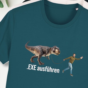 Könnte beinhalten: Ein türkisfarbenes T-Shirt mit einem Aufdruck eines Tyrannosaurus Rex, der von einer Person an der Leine geführt wird. Der Text auf dem Shirt lautet '.EXE ausführen'.