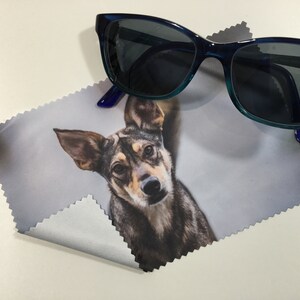 May include: A microfiber cleaning cloth with a photo of a dog printed on it. The cloth is folded in half and a pair of blue sunglasses are lying on top of it.