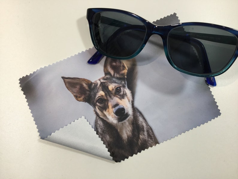 May include: A microfiber cleaning cloth with a photo of a dog printed on it. The cloth is folded in half and a pair of blue sunglasses are lying on top of it.