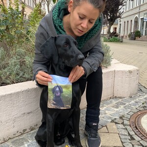 May include: A woman in a gray jacket and green scarf is holding a black dog. The dog is sitting on a stone surface and has a small fabric square with a picture of a dog on it.