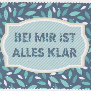 May include: A blue and white patterned card with a white border and a cream-colored label. The label has a blue and white striped background and the text "Bei mir ist alles klar" in black.