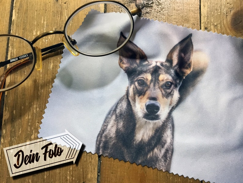 May include: A white microfiber cleaning cloth with a printed image of a dog's face. The dog has brown, black, and gray fur. The text "Dein Foto" is printed on a brown tag in the lower left corner of the image.