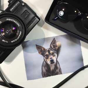 May include: A black camera with a lens labeled "PENTACON PRAKTICA 1:1.8 f=50mm MC" and a black leather strap. A black case with a pair of blue sunglasses is in the upper right corner. A white microfiber cleaning cloth with a photo of a brown and black dog printed on it is in the center of the image.