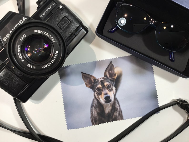 May include: A black camera with a lens labeled "PENTACON PRAKTICA 1:1.8 f=50mm MC" and a black leather strap. A black case with a pair of blue sunglasses is in the upper right corner. A white microfiber cleaning cloth with a photo of a brown and black dog printed on it is in the center of the image.