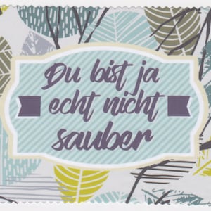 May include: A light blue card with a white border and a scalloped edge. The card has a light blue and white striped background with a pattern of leaves in shades of green, yellow, and gray. The card has a white label with a light blue border and the text "Du bist ja echt nicht sauber" in purple.