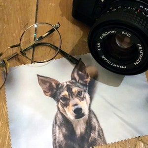 May include: A white microfiber cleaning cloth with a printed image of a brown and black dog. The dog is looking directly at the camera. The cloth is lying on a wooden surface. A pair of round glasses and a black camera lens are also visible in the image.