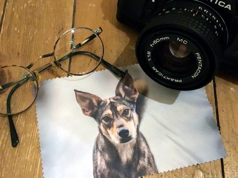 May include: A white microfiber cleaning cloth with a printed image of a brown and black dog. The dog is looking directly at the camera. The cloth is lying on a wooden surface. A pair of round glasses and a black camera lens are also visible in the image.