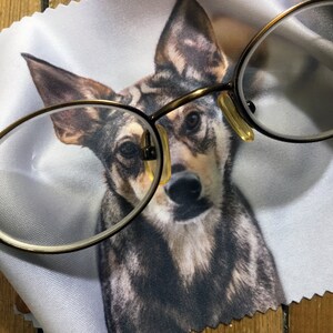 May include: A close-up of a round, brown-framed pair of glasses resting on a white microfiber cleaning cloth with a printed image of a dog's face. The dog has brown, black, and gray fur.
