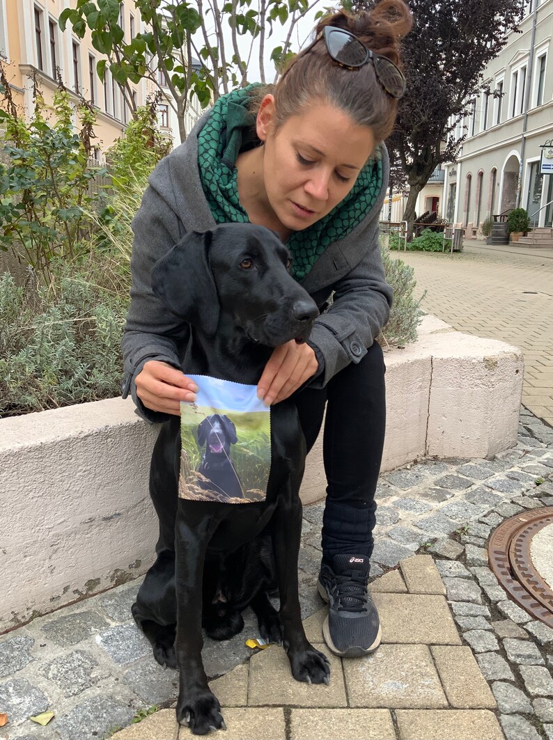 Pode incluir: Uma mulher com uma jaqueta cinza e um len&ccedil;o verde est&aacute; segurando um cachorro preto. O cachorro est&aacute; sentado em uma superf&iacute;cie de pedra e tem um pequeno quadrado de tecido com uma imagem de um cachorro nele.