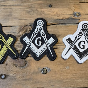 May include: Three embroidered patches featuring the Masonic Square and Compasses symbol. One patch has a black background with gold details, another with white details on black, and the third with black details on white. The patches are on a wooden surface.