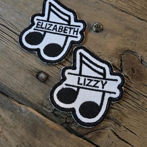 May include: Two embroidered patches in the shape of a musical note, with the names "ELIZABETH" and "LIZZY" in white lettering. The patches have a black and white color scheme and are set against a rustic wooden background.