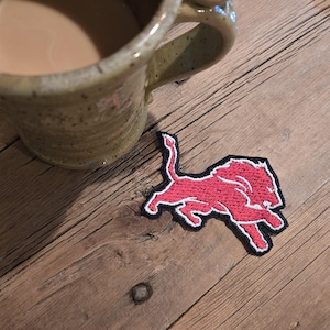 May include: A red lion patch with a black border is placed on a wooden surface next to a mug of coffee. The patch features a running lion design. The mug is a speckled brown color.