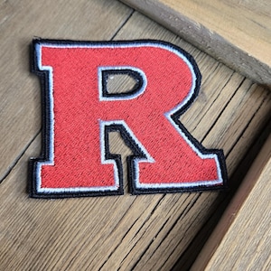 May include: Embroidered patch in the shape of the letter "R" in red with a white border and black outline. The patch is on a wooden surface. The letter is a collegiate style patch.