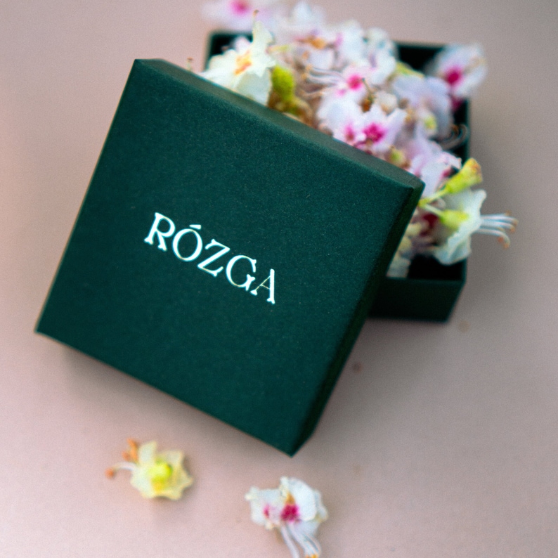 Op de afbeelding: Een groene geschenkdoos met het woord "R&Oacute;ŻGA" in wit gedrukt op de bovenkant. De doos is open en gevuld met witte en roze bloemen.