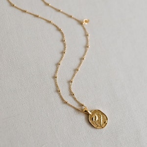 Op de afbeelding: Een gouden ketting met een delicate ketting en een hanger met een bloemenmotief. De hanger is ovaalvormig en heeft een gedetailleerd bloemenmotief.