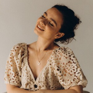 Op de afbeelding: Een vrouw draagt een wit gehaakt topje met een delicate gouden ketting en een klein hangertje. Ze glimlacht en kijkt omhoog.