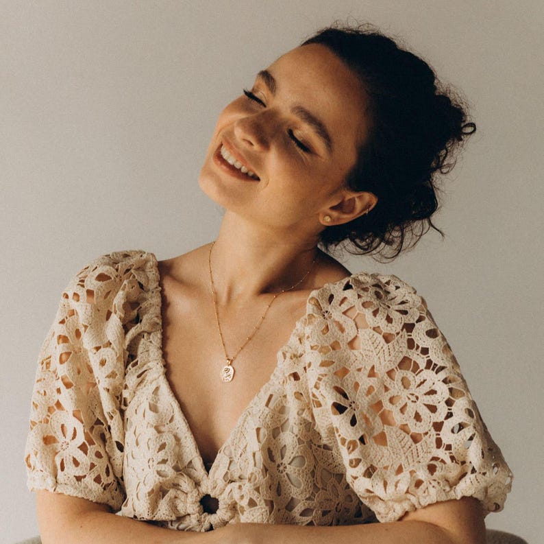 Op de afbeelding: Een vrouw draagt een wit gehaakt topje met een delicate gouden ketting en een klein hangertje. Ze glimlacht en kijkt omhoog.