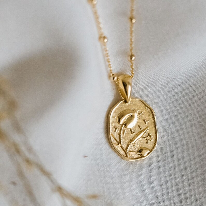 Op de afbeelding: Gouden ketting met een delicate ketting en een hanger met een gestileerd bloemmotief. De hanger is ovaalvormig en heeft de gravure "ELIA".