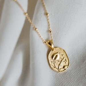 Op de afbeelding: Een gouden ketting met een delicate ketting en een hanger met een gestileerd bloemendesign. De hanger is ovaalvormig en heeft een reli&euml;fbloemmotief met de inscriptie "Tilia".