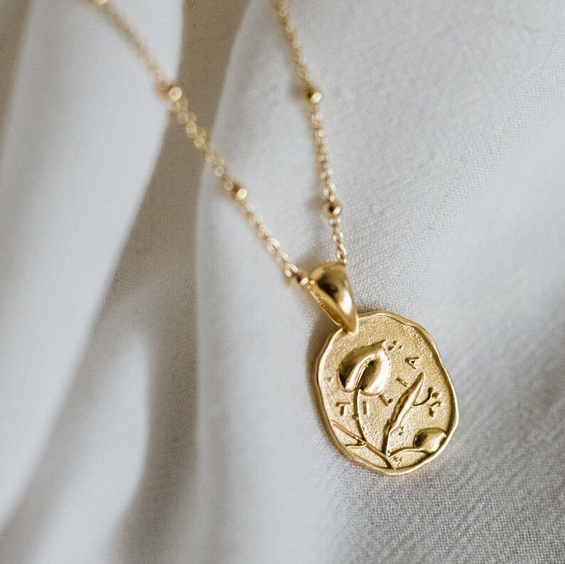 Op de afbeelding: Een gouden ketting met een delicate ketting en een hanger met een gestileerd bloemendesign. De hanger is ovaalvormig en heeft een reli&euml;fbloemmotief met de inscriptie "Tilia".