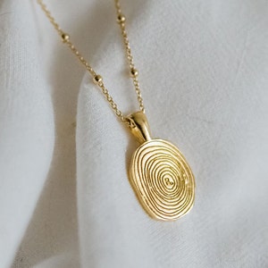 Op de afbeelding: Een gouden ketting met een ronde hanger met een concentrisch cirkelontwerp. De hanger is gemaakt van getextureerd metaal en hangt aan een delicate ketting.