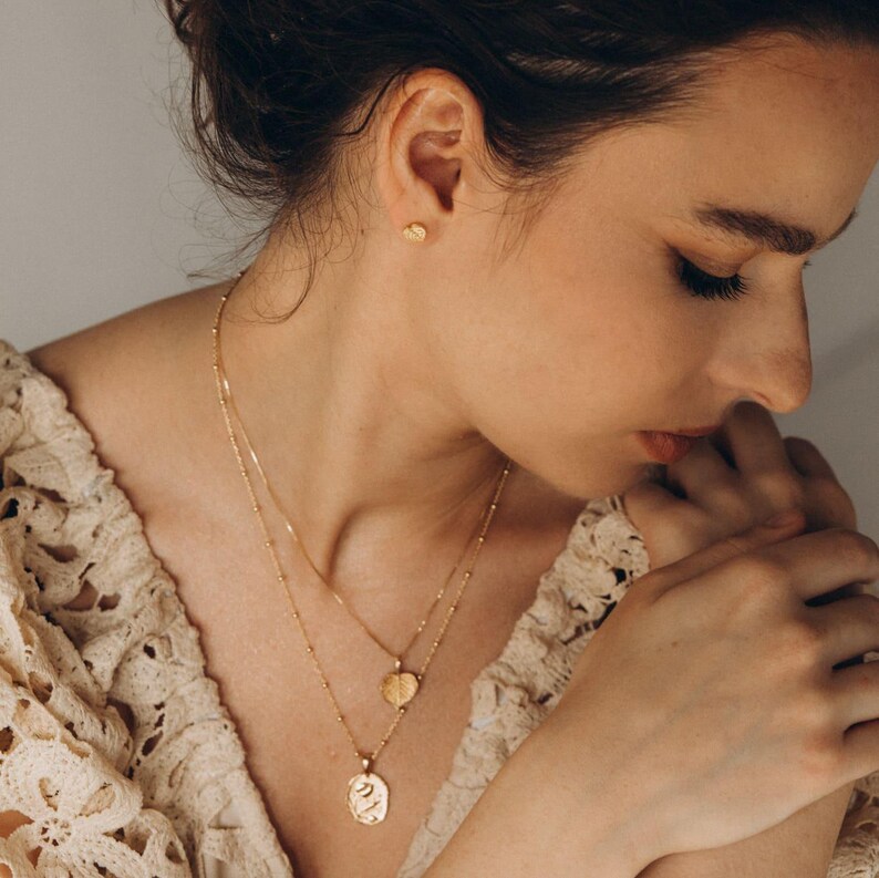 Op de afbeelding: Een vrouw draagt een gouden ketting met twee hangers. De grotere hanger is een getextureerde ovale vorm, en de kleinere hanger is een blaadje. Ze draagt ook een kleine gouden oorbel.