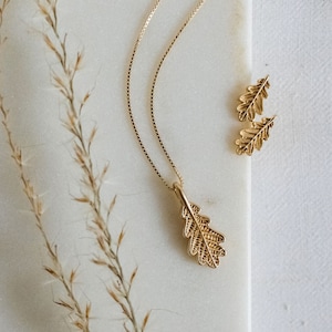May include: A gold necklace with a delicate oak leaf pendant and matching earrings. The necklace is on a white surface with dried grass.