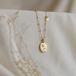 Op de afbeelding: Een gouden ketting met een delicate ketting en een hanger met een bloemenmotief. De hanger is ovaalvormig en heeft een textuur.