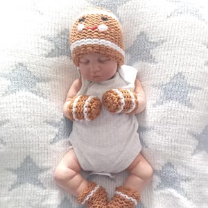 May include: A crochet set consisting of a tan hat with a gingerbread face design, two matching mittens, and two booties. Each piece features white horizontal stripes. The hat has embroidered black eyes and a red mouth. The set is worn by a doll.