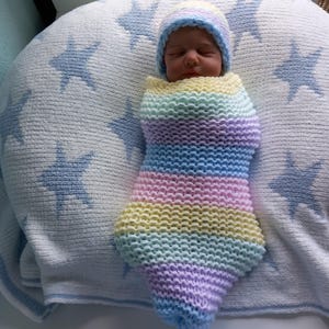 Puede incluir: Un bebé recién nacido envuelto en una manta de punto de color arcoíris pastel y un gorro a juego. El bebé descansa sobre una manta blanca con diseños de estrellas azules. La manta y el gorro son hechos a mano.