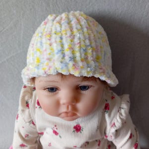 Puede incluir: Una muñeca bebé con un gorro de punto blanco multicolor con un borde pequeño. El gorro tiene motas amarillas, rosas y azules. La muñeca también lleva una camisa de manga larga color crema con un estampado floral.
