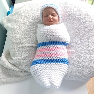 Puede incluir: Un bebé recién nacido envuelto en una envoltura de punto blanca con rayas azules y rosas. El bebé lleva un gorro blanco a juego con un ribete azul. La envoltura está sobre una manta blanca y esponjosa.