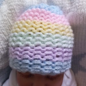 May include: A pastel rainbow-striped knitted baby hat. The hat features bands of light blue, pink, yellow, and lavender. The hat is made of soft yarn and is designed to fit a baby's head. The hat is shown on a baby.