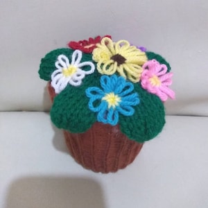 May include: A crocheted flower pot with a brown pot and green leaves. The pot is decorated with colourful flowers in shades of white, yellow, blue, pink, and red.