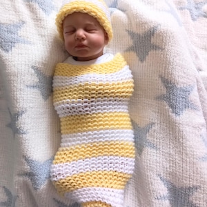 Puede incluir: Un bebé recién nacido envuelto en un capullo de punto a rayas amarillas y blancas y un gorro a juego. El bebé está acostado sobre una manta blanca con diseños de estrellas azules. Los ojos del bebé están cerrados.
