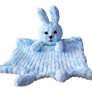 Puede incluir: Una manta para bebé de color azul claro con un conejito de peluche adjunto. El conejito tiene ojos negros y una nariz negra.