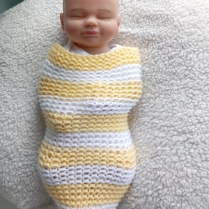 Puede incluir: Una muñeca bebé envuelta en un capullo tejido a mano con rayas amarillas y blancas. La muñeca tiene los ojos cerrados y una expresión serena. El capullo tejido está diseñado para envolver el cuerpo de la muñeca.