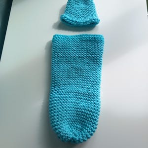 Puede incluir: Un conjunto de capullo y gorro de bebé tejido en color turquesa. El capullo tiene forma cilíndrica y el gorro es un gorrito pequeño. El conjunto está hecho de tejido de punto texturizado. Mide aproximadamente 50 cm de largo.