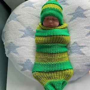 Puede incluir: Un bebé recién nacido envuelto en un disfraz de oruga tejido de color verde y amarillo, con un gorro a juego con antenas. El bebé está acostado sobre una almohada blanca con patrones de estrellas azules.