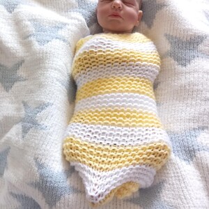 May include: A realistic baby doll wrapped in a handmade knitted yellow and white striped swaddle. The doll is lying on a soft, white blanket with blue star patterns. The baby's eyes are closed.