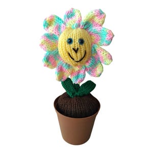 May include: A knitted flower with a smiling face in a brown pot. The flower is yellow, pink, and blue.