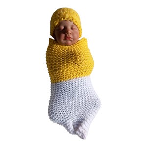 Puede incluir: Muñeca bebé recién nacida con un gorro amarillo de ganchillo y envuelta en un capullo de ganchillo amarillo y blanco. Los ojos del bebé están cerrados. Fondo blanco.