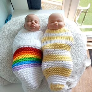 Puede incluir: Dos muñecas bebé envueltas en capullos de punto hechos a mano. Un capullo es blanco con una franja arcoíris, el otro es de rayas amarillas y blancas. Las muñecas están sobre una superficie blanca y esponjosa.