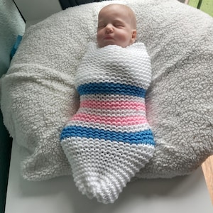 Puede incluir: Un bebé recién nacido envuelto en un capullo tejido a mano. El capullo es blanco con rayas horizontales azules y rosas. El bebé descansa sobre una almohada blanca y esponjosa.