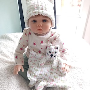 May include: A baby doll wearing a white cat-eared hat and a floral print shirt. The doll holds a matching white cat security blanket with pink and green floral accents. The doll is sitting on a white, fluffy blanket.
