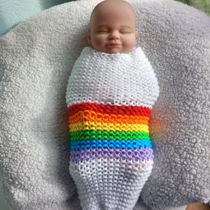 Puede incluir: Una muñeca para bebé envuelta en un capullo de punto blanco con un diseño de rayas arcoíris. La muñeca está sobre una superficie suave y esponjosa de color blanco. Los colores del arcoíris incluyen rojo, naranja, amarillo, verde, azul y morado.