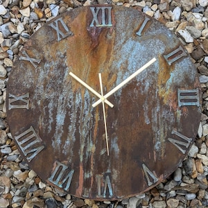 Può includere: Un orologio rotondo in metallo invecchiato con numeri romani e lancette dorate. Il quadrante dell'orologio ha un aspetto strutturato color ruggine con accenti blu-verdi. È un oggetto decorativo.